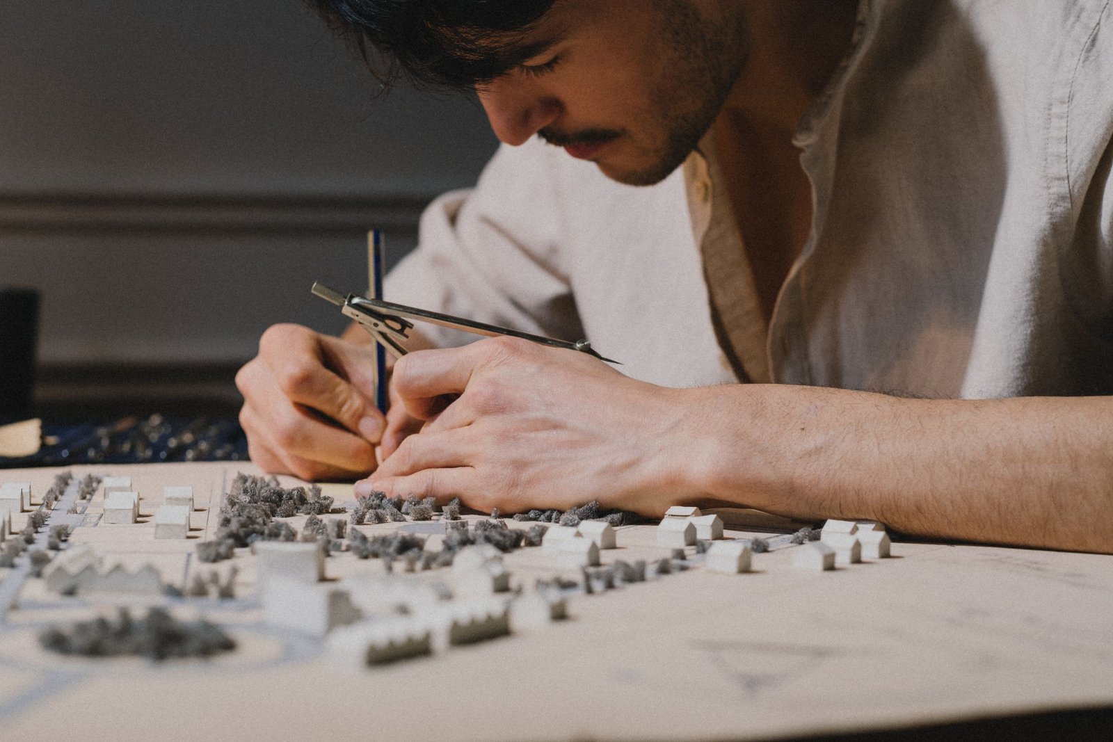A model maker leans over a miniature town of small white houses, measuring carefully with a pair of compasses