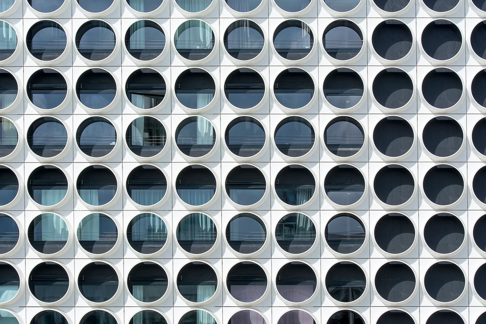 Facade of identical white square panels, each framing a round window, repeating in a precise grid with curtains varying behind the glass