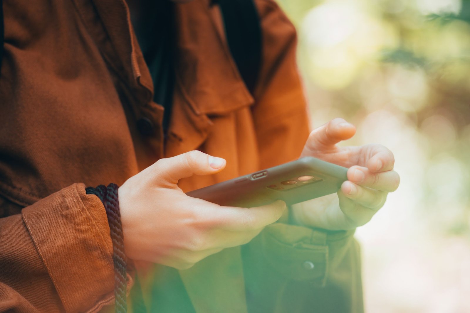Hands holding a smartphone sideways outdoors, a person in a rust jacket with soft green foliage blurred behind