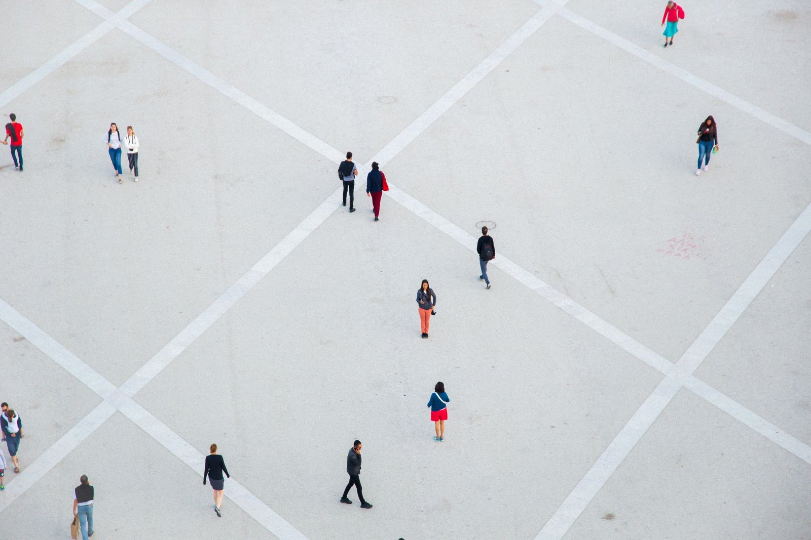 Pedestrians walking far apart across a pale paved square crossed by broad diagonal lines, seen from high above