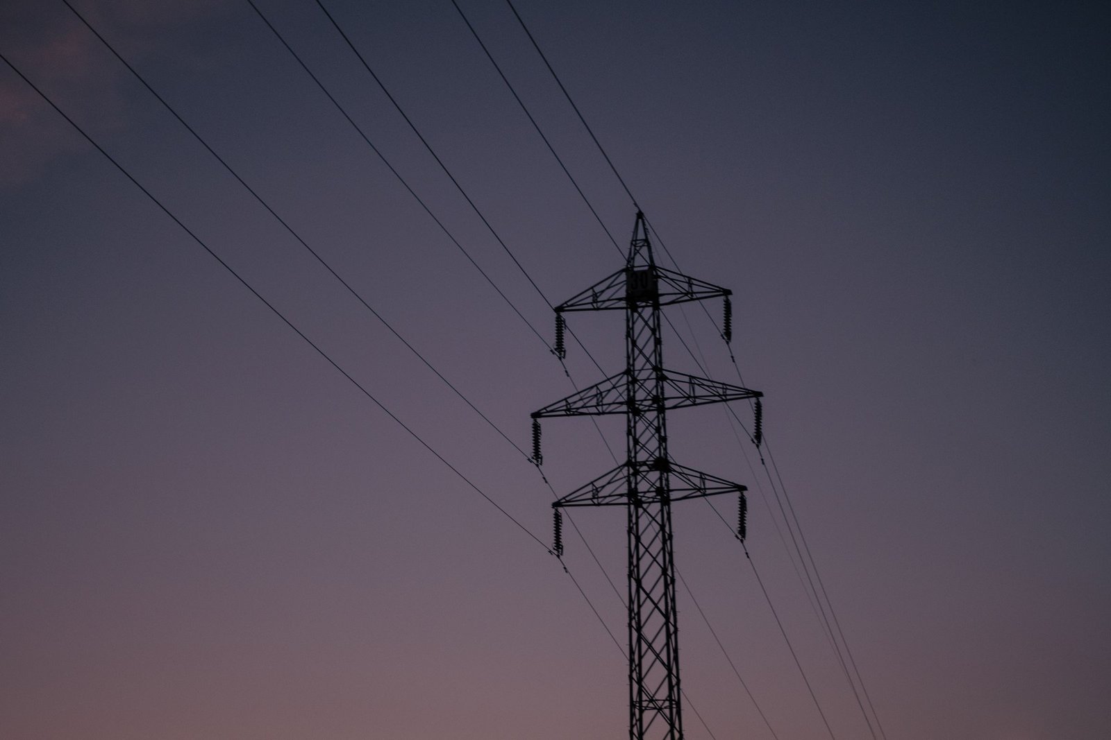 Silhouette of a lattice electricity pylon against a slate and mauve dusk sky, its power lines running diagonally across the frame