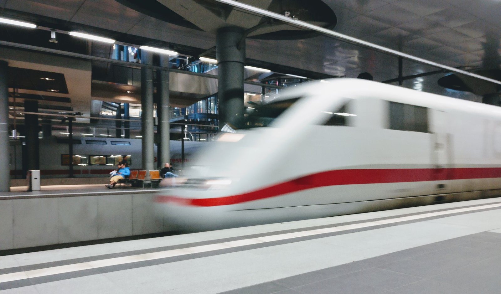 A white high-speed train blurs past an indoor station platform, its red stripe streaking beneath grey columns and a glossy ceiling