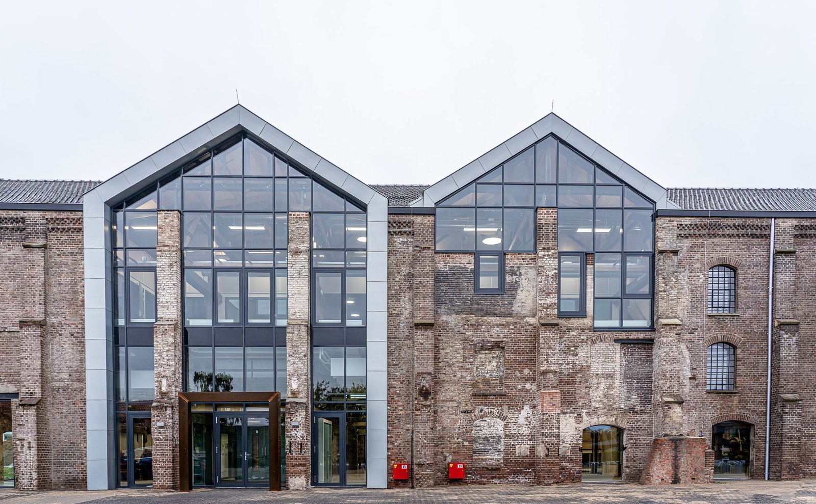 Weathered brick industrial building renovated with two tall glazed gables, the old walls kept around a modern glass entrance