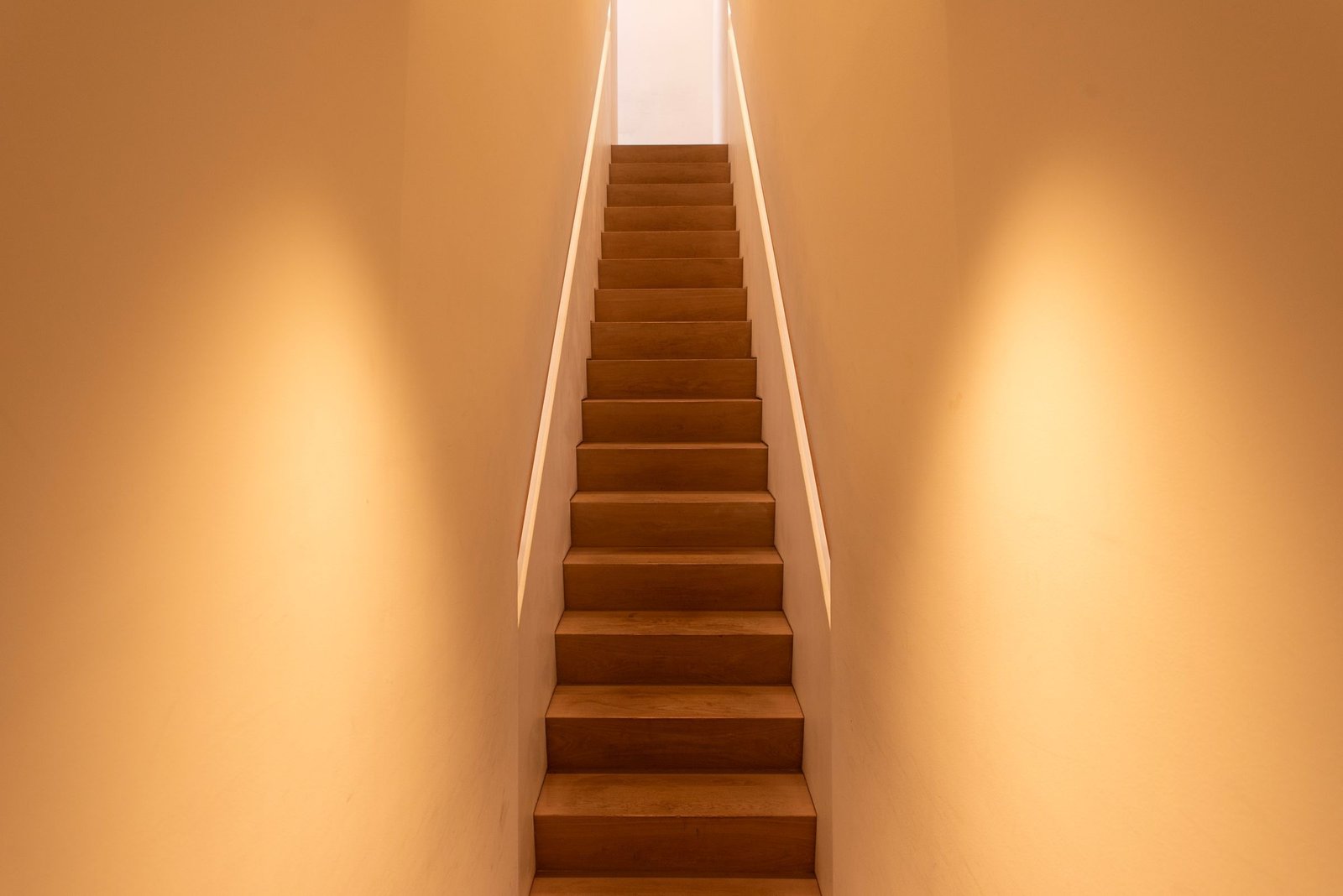 A narrow wooden staircase climbing between warmly lit plaster walls towards a bright opening at the top