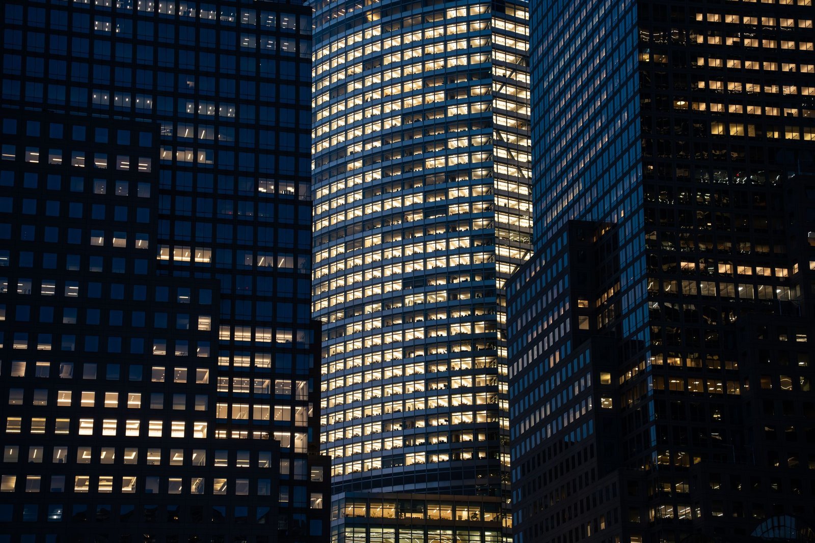 A glass office tower glowing floor by floor at dusk, its lit windows standing out between darker neighbouring towers