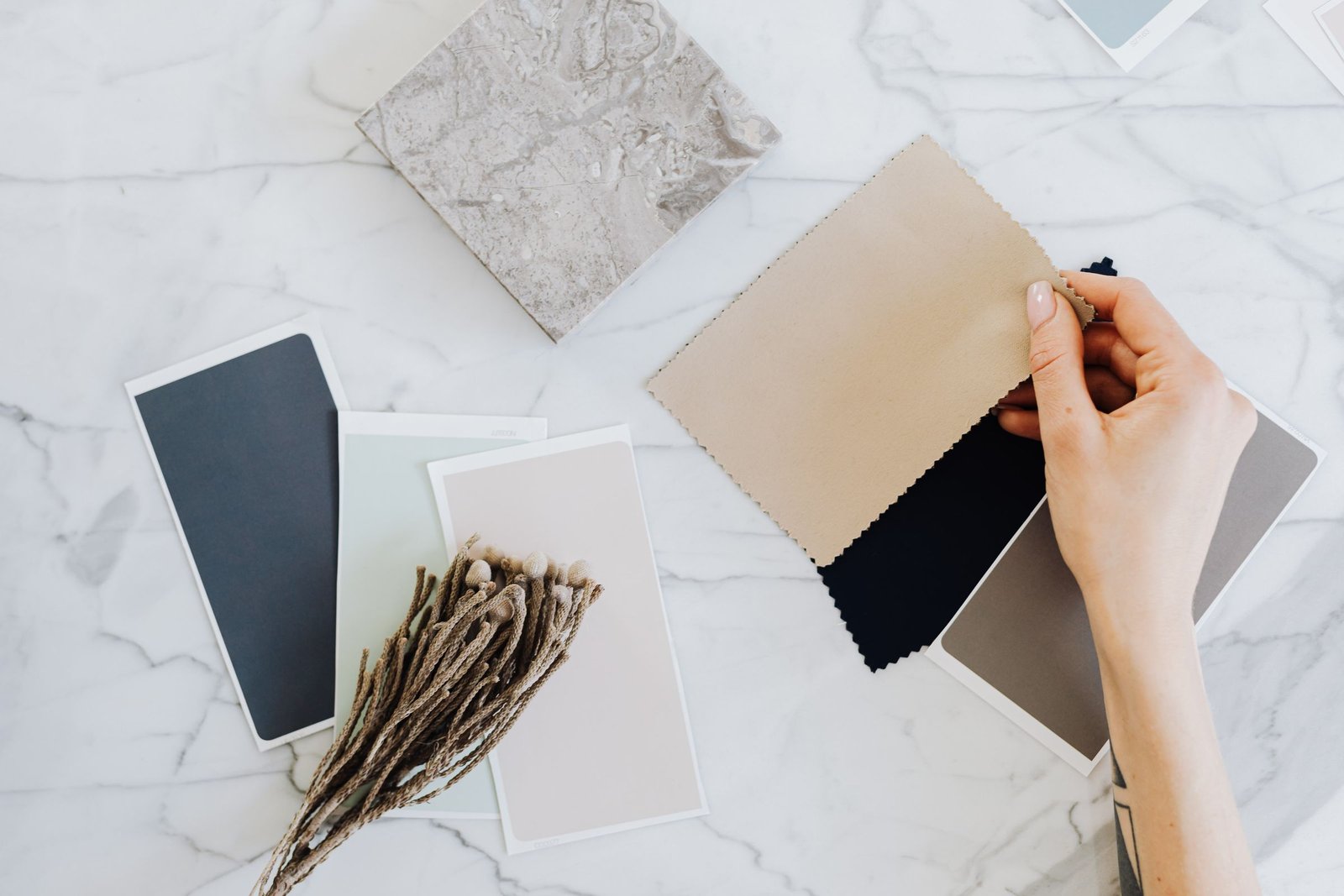 Hand holding a beige fabric swatch beside slate, sage and blush paint cards, a stone tile and dried stems on a white marble table