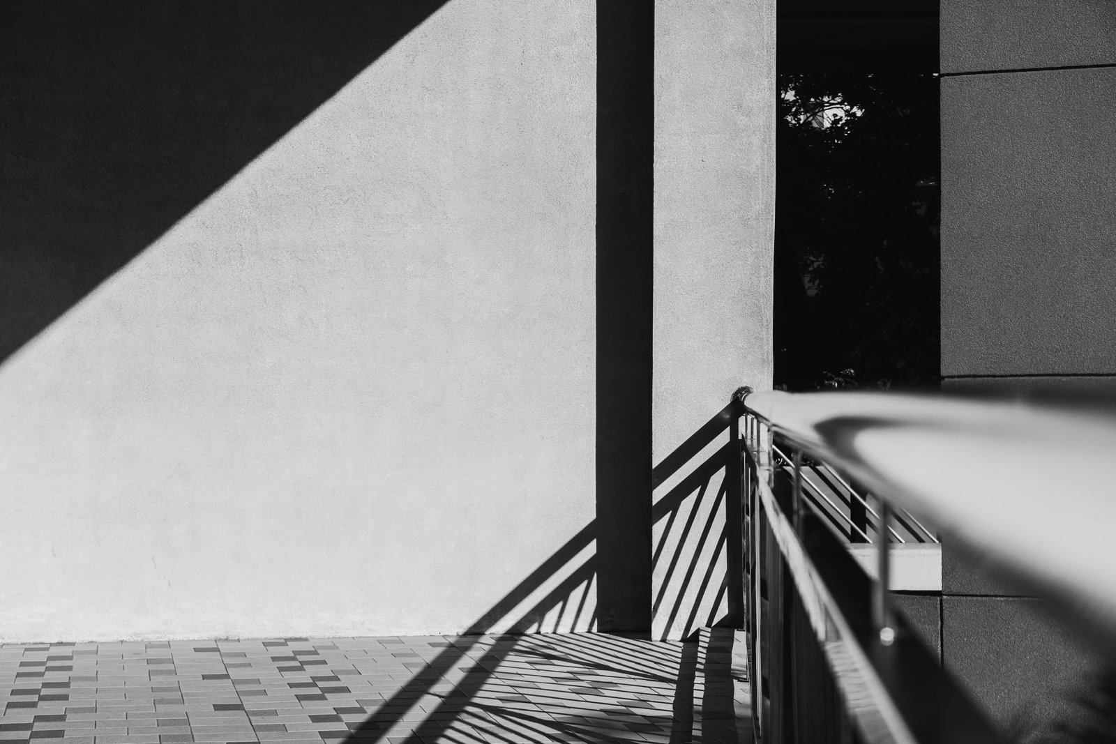 Handrail beside a plain rendered wall in black and white, its railings casting crisp diagonal shadows that lead towards a shaded opening