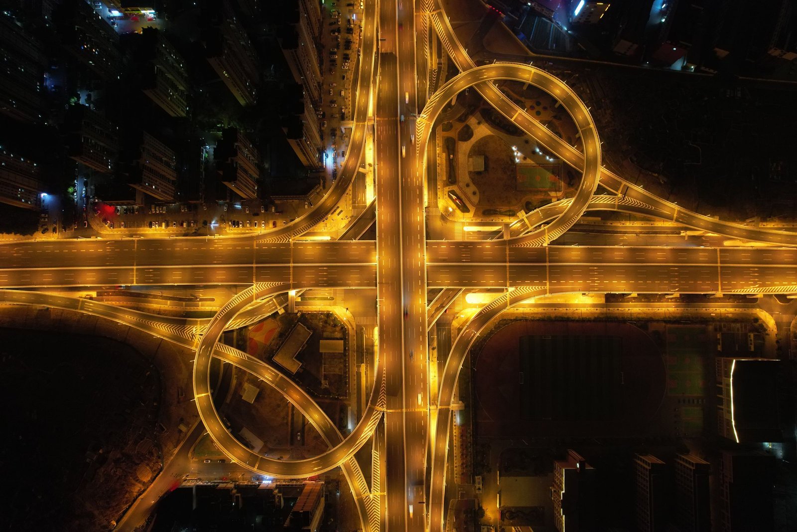 A road interchange at night seen from directly above, its glowing looping ramps joining two crossing carriageways