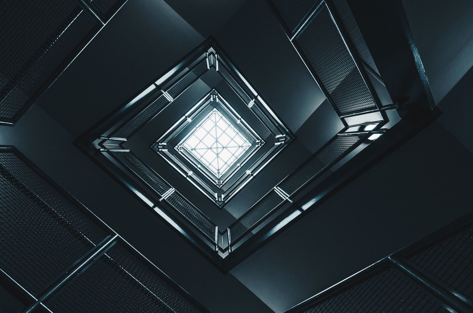 Looking up a dark square stairwell, flights of steps and railings narrowing towards a single bright skylight at the top