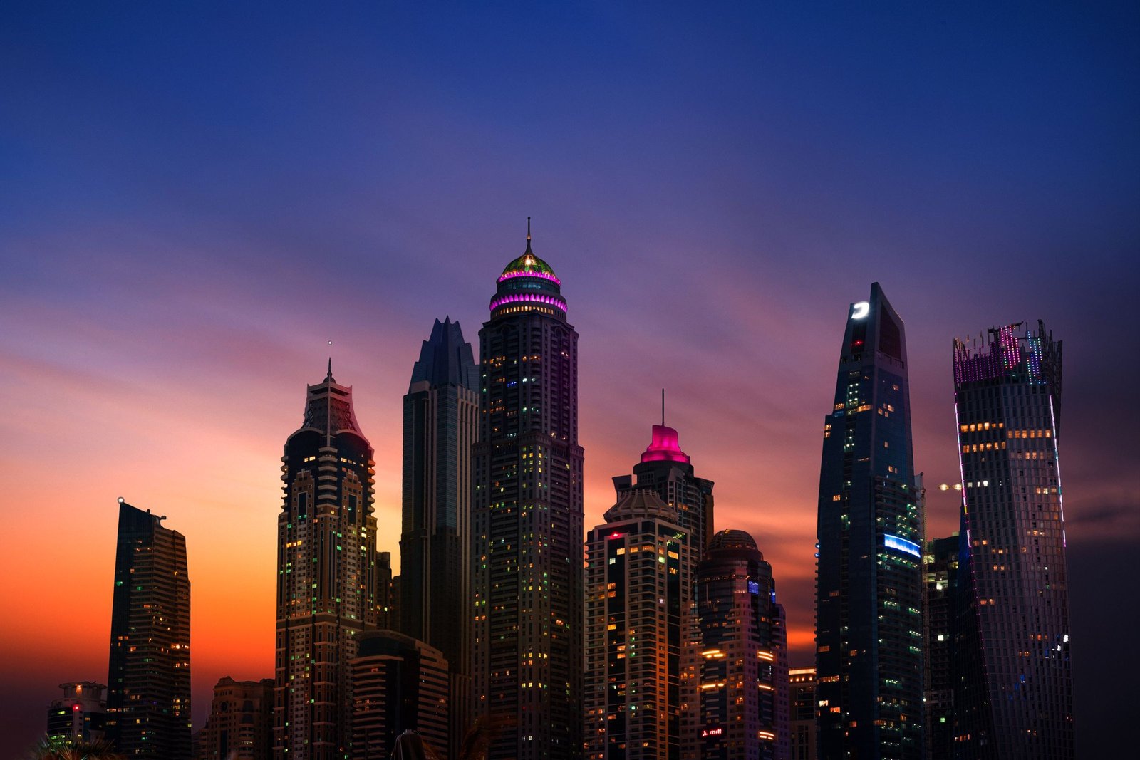 Glass towers silhouetted against a violet and amber dusk sky, their windows and crowns lit in soft pink and purple