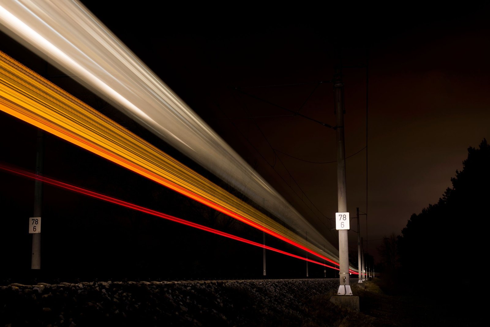 Long exposure of a train at night, its lights drawn into smooth white, amber and red streaks past a line of trackside poles