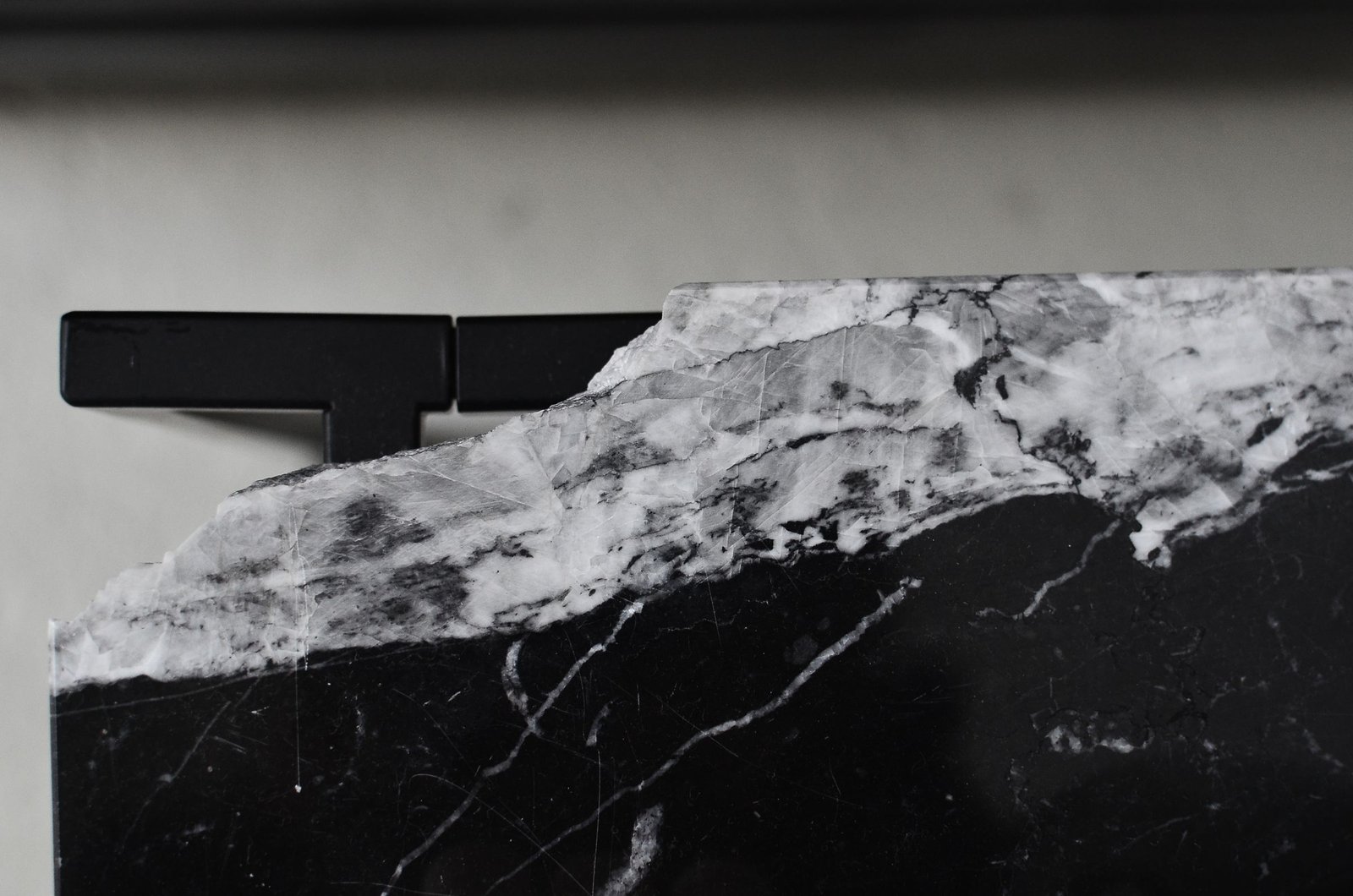 Black marble slab veined with white, its raw crystalline top edge meeting a precise matte black metal fixture in soft grey light
