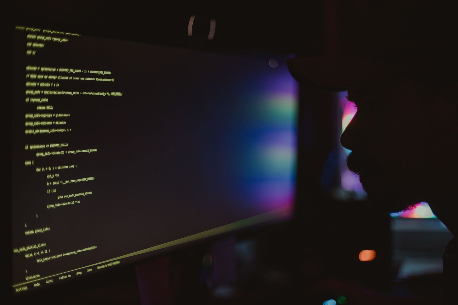 A developer seen in silhouette against violet and green light studies lines of code on a wide monitor in near darkness
