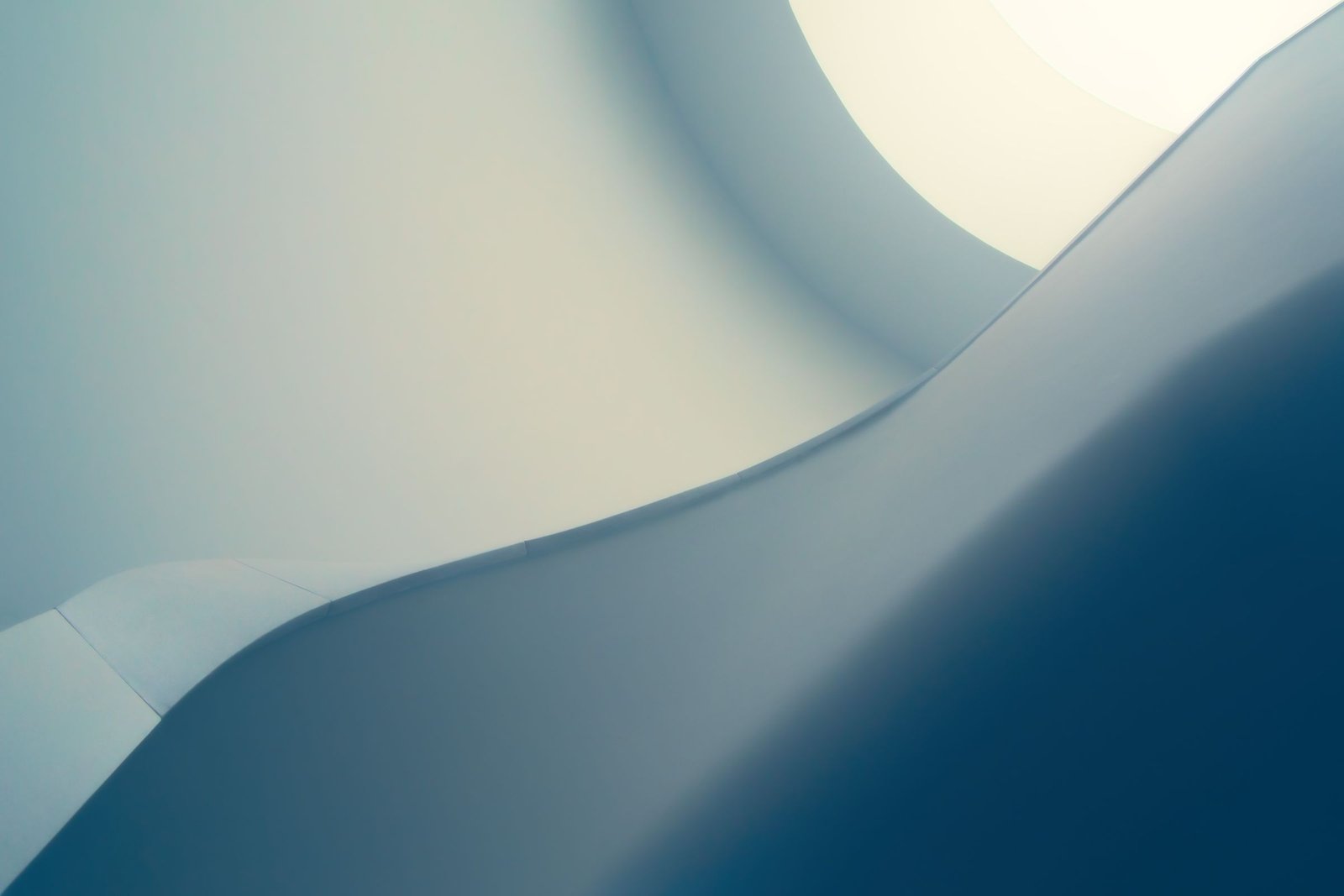 Underside of a curving white staircase seen from below, its smooth edge rising through soft blue shadow towards bright daylight